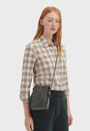 Young woman with red hair wearing a beige checkered shirt and black crossbody leather bag, standing against a plain background.
