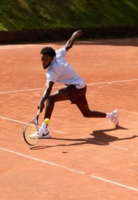 Athletisk mandlig spiller iført en lys blå skjorte og maroon shorts, som slår en gul tennisbold med en ketsjer på en grusbane.