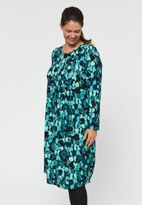 Dlhý rukáv šaty v teal a modrej farbe s kruhovým vzorom, okrúhlym výstrihom a voľným strihom, vyrobené z jemnej látky s miernym leskom.