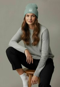 Light green ribbed beanie with logo, gray sweater, black pants, and white socks. Model sitting on wooden stool. Soft textures and neutral colors.