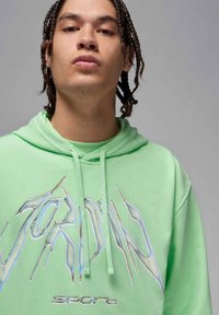 Felpa verde realizzata in morbido tessuto presenta un cappuccio, cordini e un grande logo stilizzato "JORDAN" con accenti iridescenti.