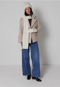 Hellrosa Wollmischjacke mit schwarzen Knöpfen, kombiniert mit einem weißen, fellen Schal, Handschuhen, einer gestrickten Mütze und blauen, weit geschnittenen Jeans. Schwarze Stiefeletten.