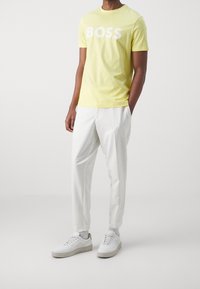 Male wearing a yellow T-shirt with "BOSS" text, white tailored pants, and white sneakers with gray socks standing against a plain background.