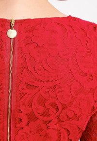 Röd spetsklänning med blommönster, avslutad med en guldfärgad dragkedja bak och strukturerade detaljer i tyget.