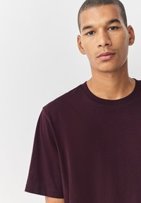 Maglietta in cotone color bordeaux con collo rotondo, maniche corte e tessuto liscio e morbido. Design semplice, senza motivi o grafiche visibili.