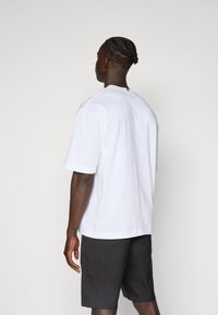 Hvid kortærmet t-shirt med afslappet pasform, lavet af bomuldsmateriale og med rund hals. Kombineret med mørke shorts, set fra ryggen.