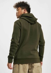 Oliven grønn hettegenser med kengurulomme, ribbestrikkede mansjetter og snor i hetten. Myk stofftekstur, enkelt design uten dekorasjoner.