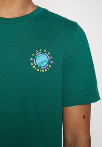 Zelené tričko s barevným kulatým logem Adidas Originals na hrudi, které nosí osoba s viditelnou pigmentovou skvrnou na paži.