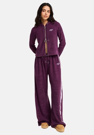 Femme debout, les mains dans le dos, portant un sweat à capuche violet zippé et un pantalon large assorti avec des bandes latérales ainsi que des chaussures compensées couleur beige.