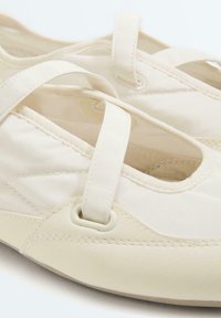 Chaussures plates blanc cassé avec une bride sur le dessus, bout rond et détails de couture visibles sur matériau synthétique.