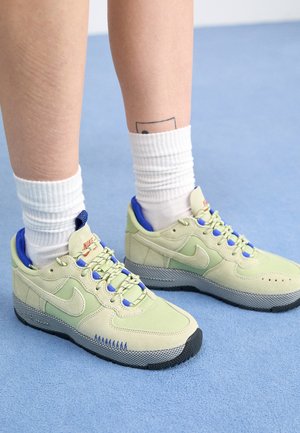 Personne portant des baskets Nike vert clair avec des touches de bleu et d'orange ainsi que des chaussettes blanches sur un tapis bleu clair.