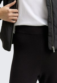 Persona che indossa un giubbotto trapuntato nero, una camicia bianca e pantaloni neri a vita alta, con la mano sinistra che tiene la cerniera del giubbotto.