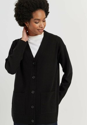 Vrouw met kort krullend haar die een zwarte gebuttonde cardigan over een wit topje draagt, kijkt naar beneden met één hand die haar nek aanraakt.