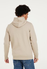 Beige Kapuzenpullover aus weichem Stoff mit einer Kängurutasche; gerippte Bündchen und Saum. Getragen mit blauen Jeans, Rückansicht.