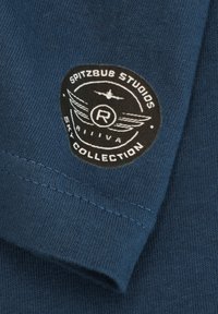 Runder schwarzer Patch mit weißem Flugzeug, Flügeln und dem Text "Spitzbub Studios Sky Collection" auf einem blauen Stoffärmel.