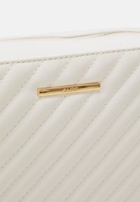 Bílá prošívaná psaníčko z umělé kůže s horizontálně žebrovanou texturou, zdobené logem zlaté barvy z kovu. Horní zapínání na zip.