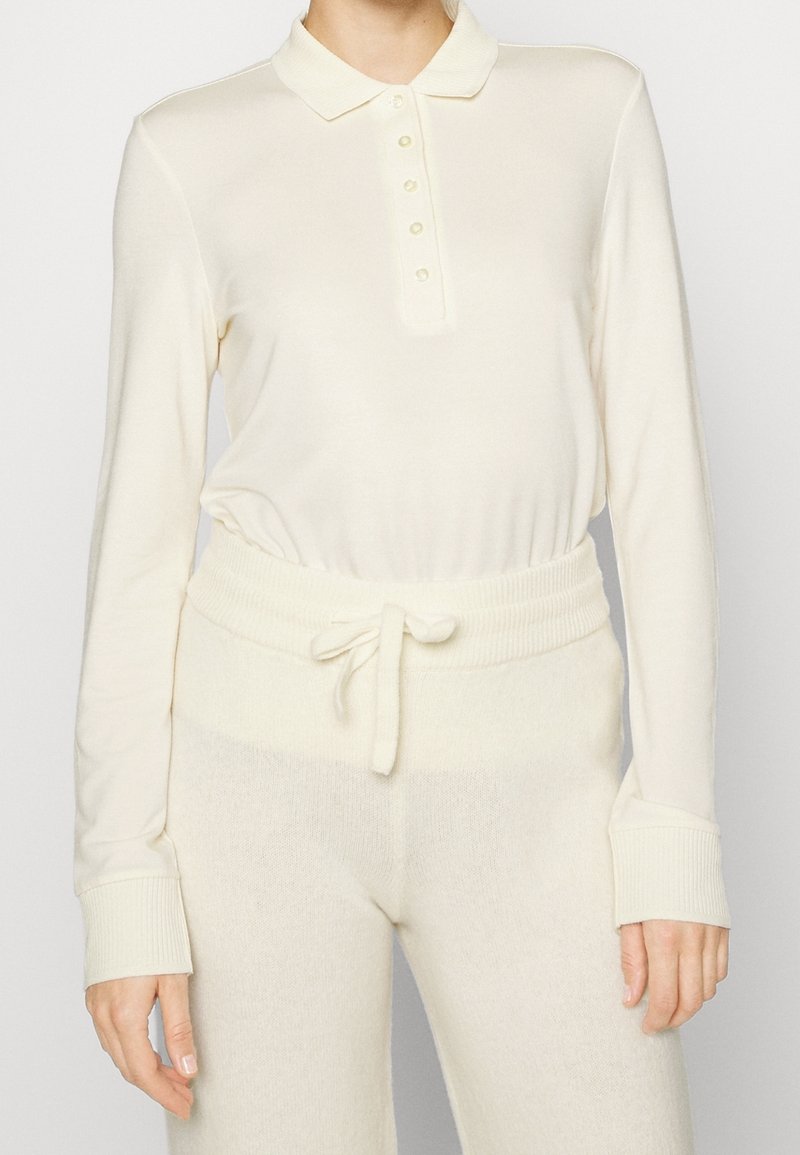 Personne portant un polo crème à manches longues avec boutons et un pantalon en maille assorti avec une taille à cordon.