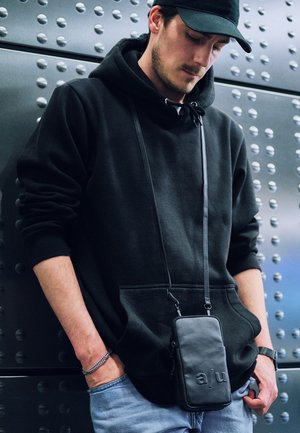 Black leather crossbody bag with embossed letters, worn over a black hoodie. Soft fabric, casual fit, and jeans with a light wash.