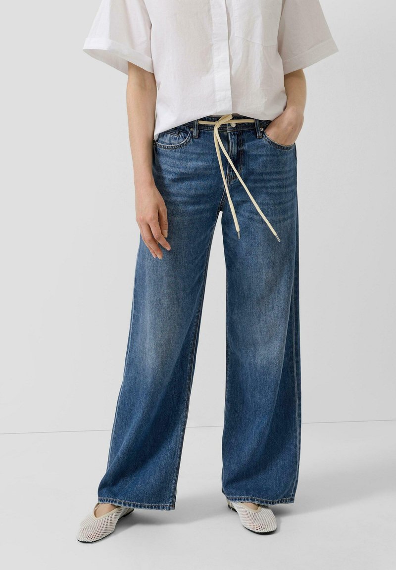 Osoba ubrana w niebieskie dzwony z białym sznurkiem, białą koszulę na krótki rękaw z guzikami oraz białe siateczkowe buty, stojąca na neutralnym tle.