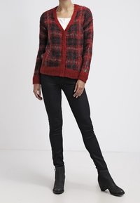 Cárdigan de punto a cuadros rojo y negro con cuello en V, mangas largas y puños acanalados, combinado con jeans skinny negros y botas hasta el tobillo.