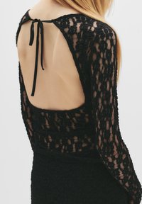 Abito in pizzo nero con maniche lunghe, schiena scoperta con chiusura a fiocco, tessuto a trama e silhouette aderente.