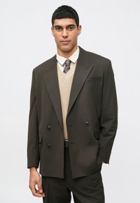 Καφέ διπλής όψης σακάκι πάνω από ένα μπεζ πλεκτό πουλόβερ και λευκό πουκάμισο, συνδυασμένο με μια σχεδιασμένη γραβάτα. Λείος ύφασμα, κομψή εφαρμογή.