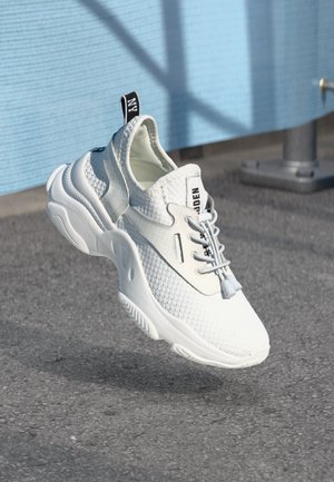 Baskets de sport blanches en tissu en maille, avec une semelle épaisse et une languette noire, flottant au-dessus d'un asphalte près d'un mur bleu.