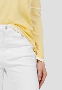 Keltainen ribattu pitkähihaine paita valkoisilla yksityiskohdilla, yhdistetty valkoisiin kapeisiin farkkuihin. Paita on rento malli ja siinä on käänteet hihoissa.