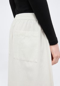 Άτομο που φοράει μαύρο μακρυμάνικο τοπ και ψηλόμεση ανοιχτό μπεζ φούστα με ελαστική μέση και μεγάλη πίσω τσέπη.