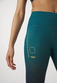 Leggings attillati color teal realizzati in tessuto elasticizzato, dotati di logo Venum giallo, scala di misurazione e motivo a rete per la traspirabilità.