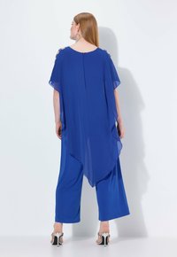 Jumpsuit bleu royal avec un design ample et fluide, superposition transparente et accents détaillés aux épaules. Fermeture éclair dans le dos et pantalons à jambes larges.