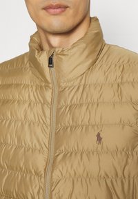 Ljusbrun quiltad pufferjacka med hög krage och frontdragkedja. Har en broderad logotyp på bröstet. Slät nylonmaterial.