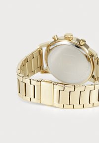 Montre bracelet en métal doré avec des maillons, trois boutons-poussoirs et logo gravé sur le fermoir, présentée de dos sur fond blanc.