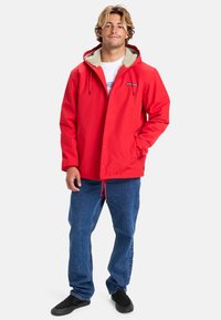 Rød vandtæt jakke med fleece-fôr i hætten, trykknapper og justerbar snor i livet. Sammen med blå jeans og sorte sko.