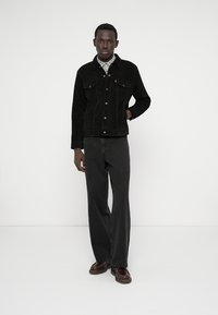 Svart mockajacka med framfickor, kombinerad med mörka svarta jeans. Modellen står mot en vit bakgrund med avslappnade händer. Bruna skor fulländar outfiten.