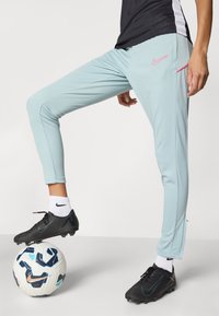 Nike light blue athletic pants with a slim fit, featuring pink accents, sporty seams, and an ankle zipper, paired with black cleats on a soccer ball.