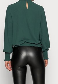 Grön blus med hög krage och detaljer med nyckelhål på baksidan, ihopparad med tighta svarta läderbyxor. Blusen har smockade manschetter och fåll.