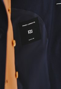 Wewnątrz czarnej marynarki znajduje się gładka granatowa podszewka z prostokątną metką z napisem "PAKO LORENTE" oraz "BLACK LINE" w białym kolorze.