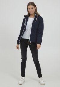 Marineblå lett jakke med hvit glidelås og snor, med hette og elastiske mansjetter, båret over en hvit skjorte og svarte jeans.