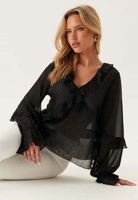 Černá průhledná blůza s volánkovým designem, dlouhými rukávy a hlubokým výstřihem do V. Vyrobena z lehkého, splývavého materiálu pro elegantní vzhled.