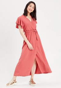 Robe maxi en corail avec des manches courtes à volants, design drapé, ceinture à la taille et fente à l'avant ; fabriquée en tissu texturé. Portée avec des sandales métalliques.