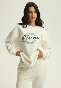 Biała bluza z czarnym napisem "Bloonii" oraz wzorem logo na środku. Połączona z dopasowanymi białymi spodniami, które cechują się miękkim materiałem i luźnym krojem.