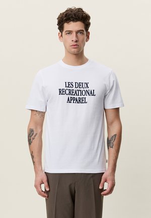Jeune homme aux cheveux bouclés portant un T-shirt blanc avec le texte "Les Deux Recreational Apparel", un pantalon marron et des tatouages visibles sur les bras, se tient face à l'avant.