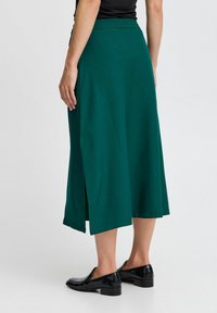 Jupe midi teal avec une taille élastique, dotée de fentes latérales. Portée avec des chaussures en cuir verni noir, mettant en valeur une texture lisse.