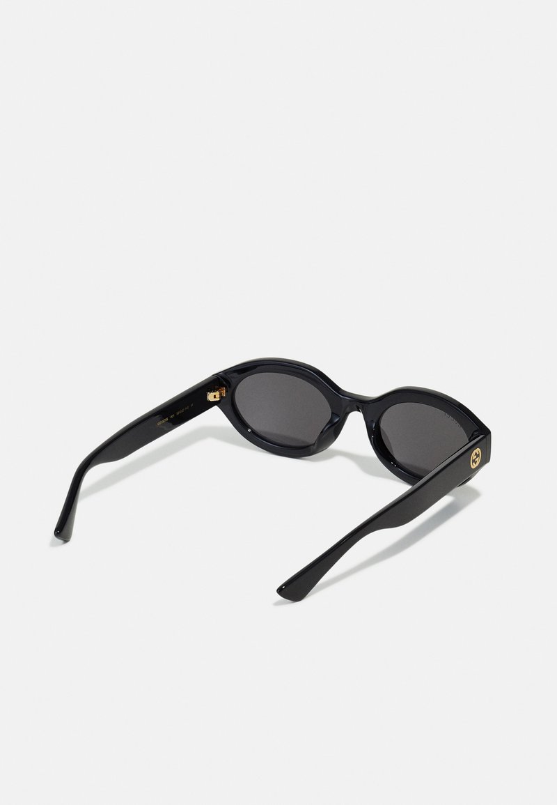 Black sunglasses with an oval lens shape, made of glossy plastic. Features gold-tone hardware and tapered black arms. Dark tinted lenses.
