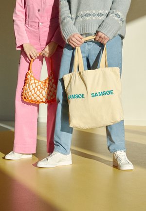 Dwie osoby stoją obok siebie, trzymając torby; jedna ma na sobie różowe spodnie i trzyma pomarańczową siatkę, druga nosi niebieskie dżinsy i trzyma beżową torbę na ramię z napisem „SAMSØE”.