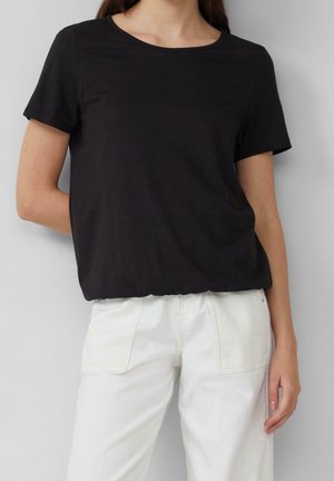 Vrouw draagt een effen zwart T-shirt met korte mouwen en witte broek, staande tegen een lichtgrijze achtergrond.