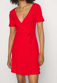 Czerwona sukienka z prążkowanego materiału o głębokim dekolcie w kształcie V, krótkich rękawach, z fasonem kopertowym oraz wiązaniem w talii. Rozkloszowany dół z gładkiej tkaniny.
