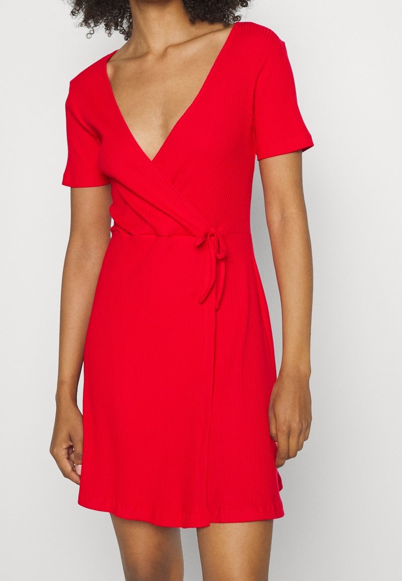 Czerwona sukienka z prążkowanego materiału o głębokim dekolcie w kształcie V, krótkich rękawach, z fasonem kopertowym oraz wiązaniem w talii. Rozkloszowany dół z gładkiej tkaniny.