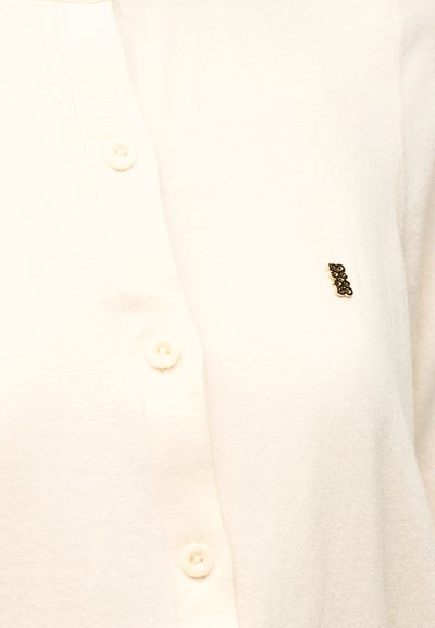 Κοντινό πλάνο ενός πουκαμίσου σε κρεμ απόχρωση με μικρό μαύρο και χρυσό έμβλημα στο στήθος κοντά στα κουμπιά.